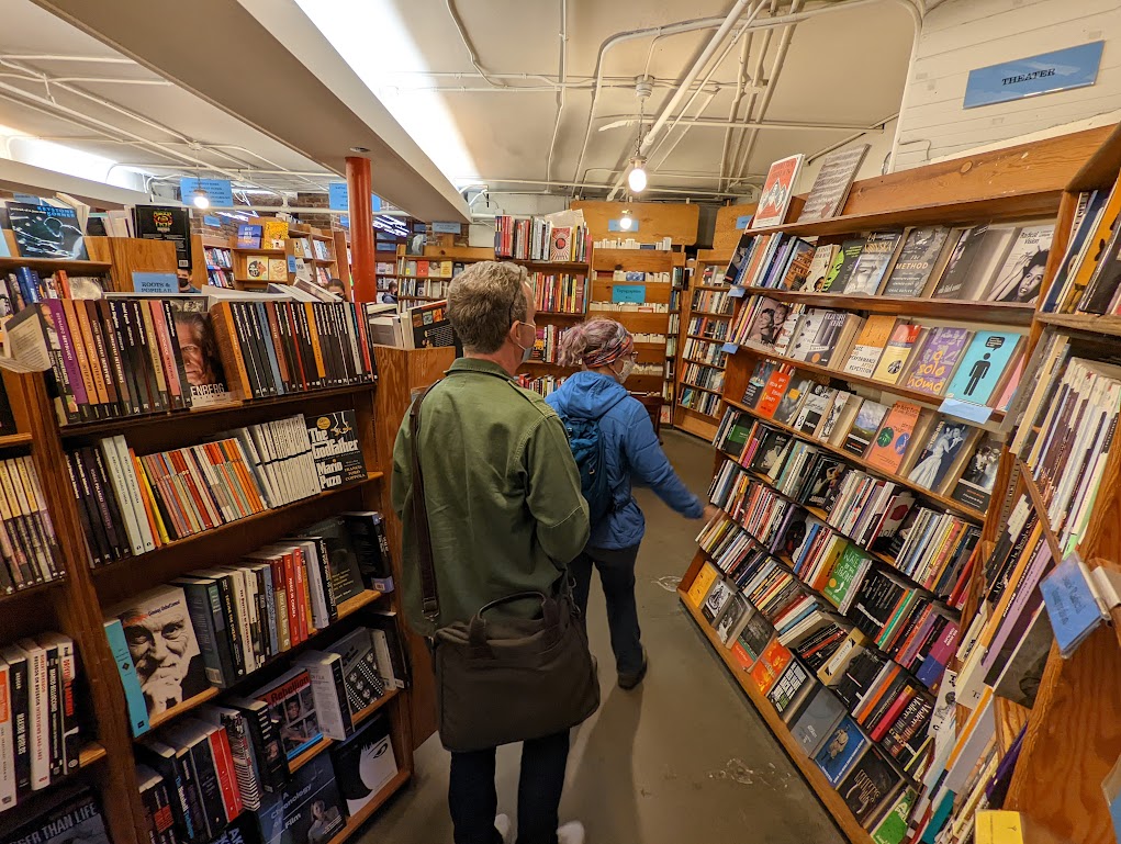 Laura and Matt walk between shelves inside the bookstore. Laura's hand is stretched out as if she wants to pet the books. She does.