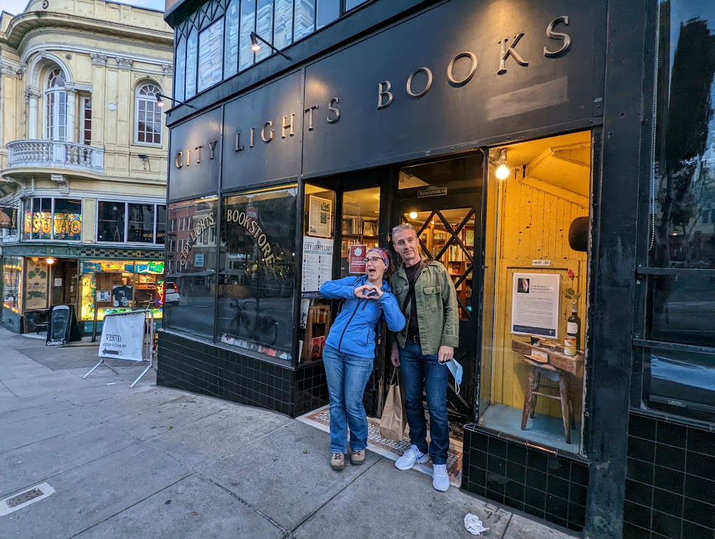 Laura and Matt stand outside the entrance of City Lights Books, leaning toward each other. Laura is holding her hands up to make a heart shape, and is making a dopey "my followers are going to love this" face. Matt appears bemused.