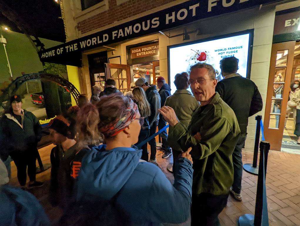 Laura, with her back to the camera, debates Matt, facing the camera, about the merits of ice cream topping while they stand in a long line in front of the Ghirardelli ice cream shop. It is night but well lit.
