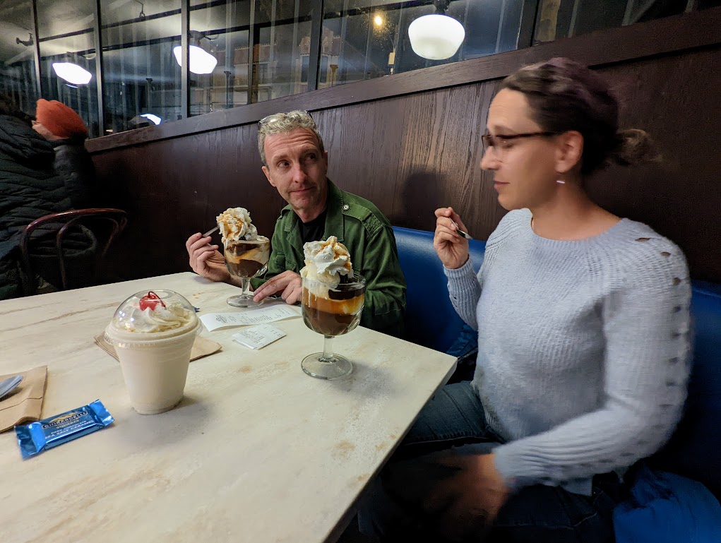 Matt and Laura sit in a blue vinyl booth with giant ice cream sundaes in front of them. The malt that presumably belongs to the photographer (Dustin) sits in the foreground. Matt is looking doubtfully over at Laura, who appears blissed out but blurry.