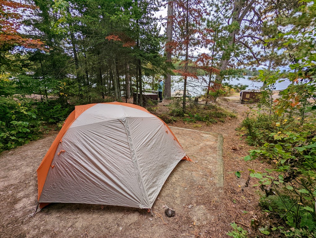 In the foreground at the lower left, a gray and orange tent is pitched on a flat, sandy surface. The sandy surface is surrounded by thin evergreen trees. Between the trees, a bit of lake can ben seen in the background.