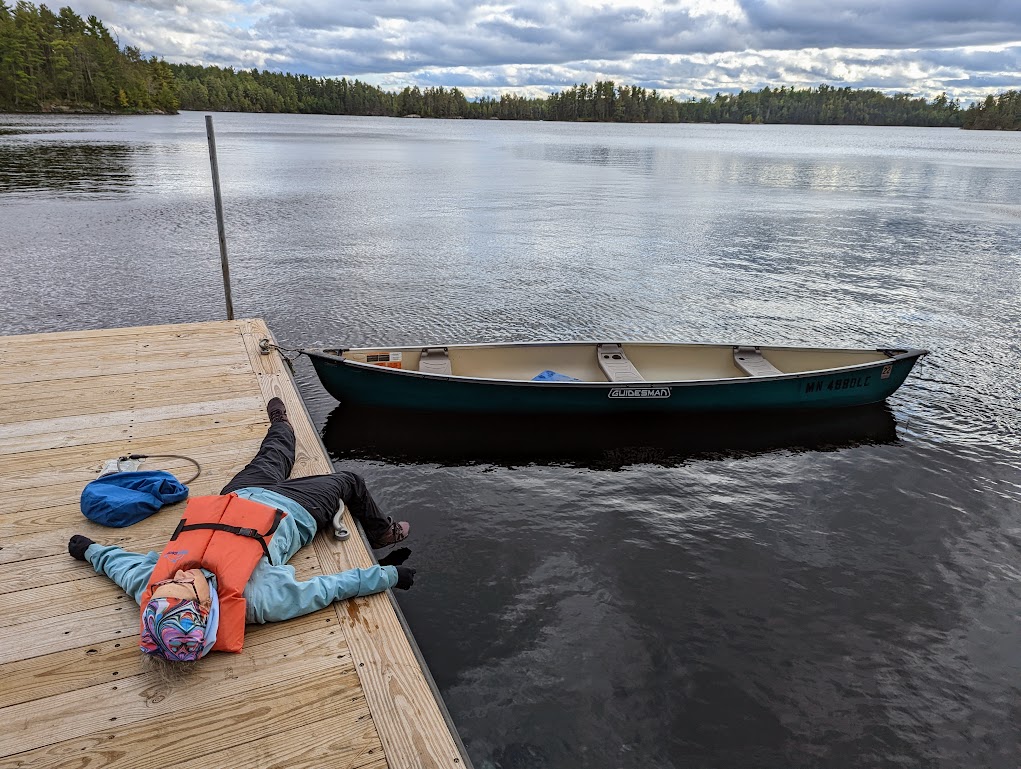 The photo is mostly water and sky. The sky is mostly cloudy with a few spots of blue. The lake is the color of steel. At the left, a wooden dock extends into the water. A green canoe is tied to its end. Laura is laying on the dock beside the canoe, face-up, one leg hanging over the side of the dock. 