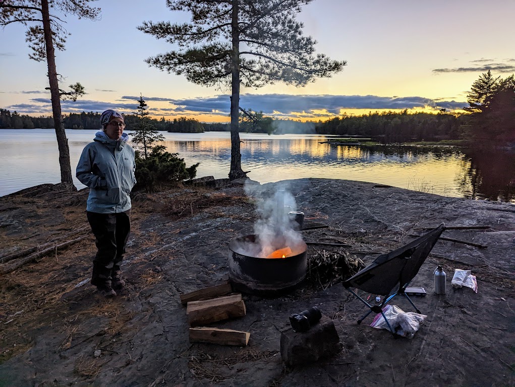 A photo of the same fire ring as above, though taken from farther back. Laura stands to the left of the fire, hands in her pockets. The lake in the background glows with the last of a sunset, with several clouds just above the horizon.