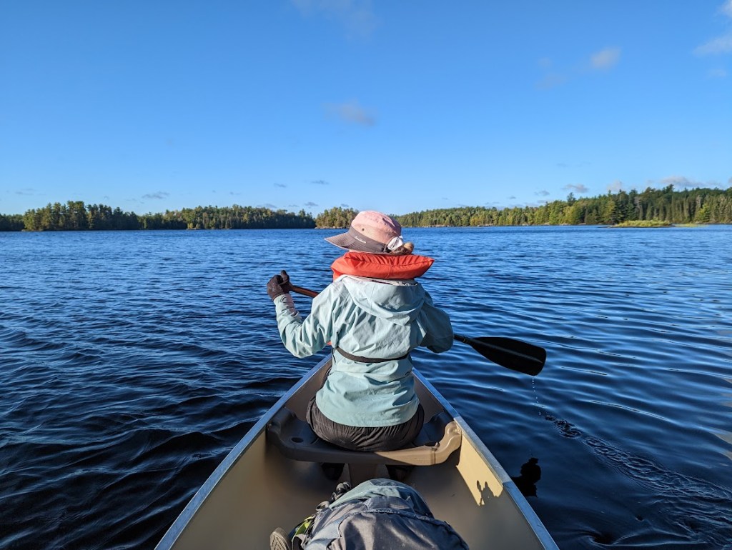 A photo taken from the back of the canoe showing Laura rowing in the front. The lake water is very, very blue, reflecting the clear sky above. 