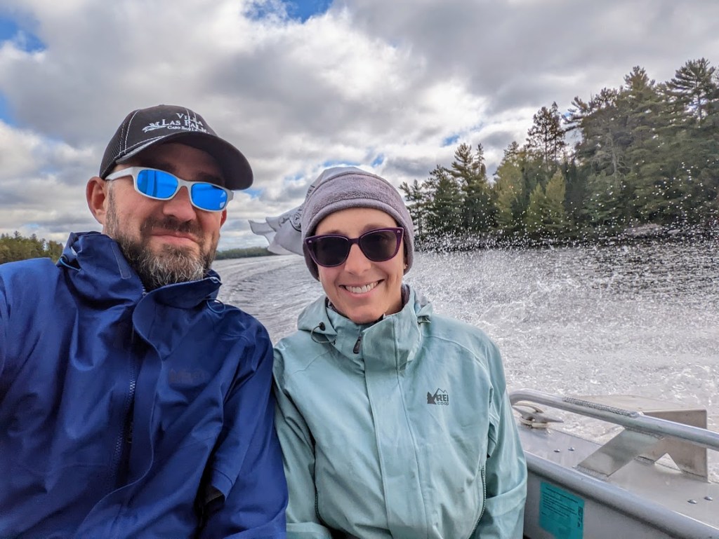 A selfie of Dustin and Laura wearing raincoats and sunglasses, smiling, in the back of the Kettle Falls Hotel's shuttle boat. Spray from the motor fans out behind them across the silver colored lake water. Trees are visible along the shore to the photo's right. A blue sky above is mostly obscured by puffy clouds. 