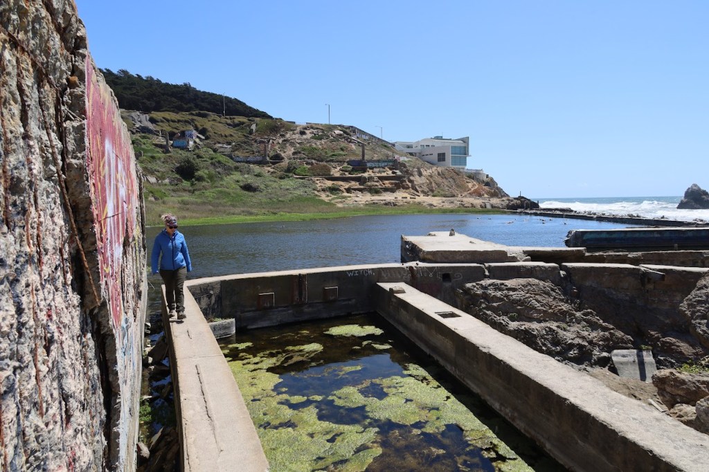 Laura walks toward the camera along a cement foundation wall with water behind her and to her right. Pond scum grows on the water to her right. The ocean can be seen in the distance.