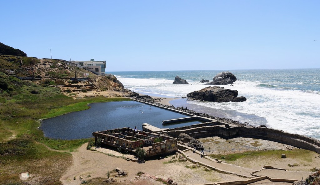 A landscape photo taken from a bluff along the Pacific Ocean. In the foreground, well below the vantage point, a pool of water is retained in a cove by a man-made wall. Additional foundation ruins can be seen to the right of the pool. 