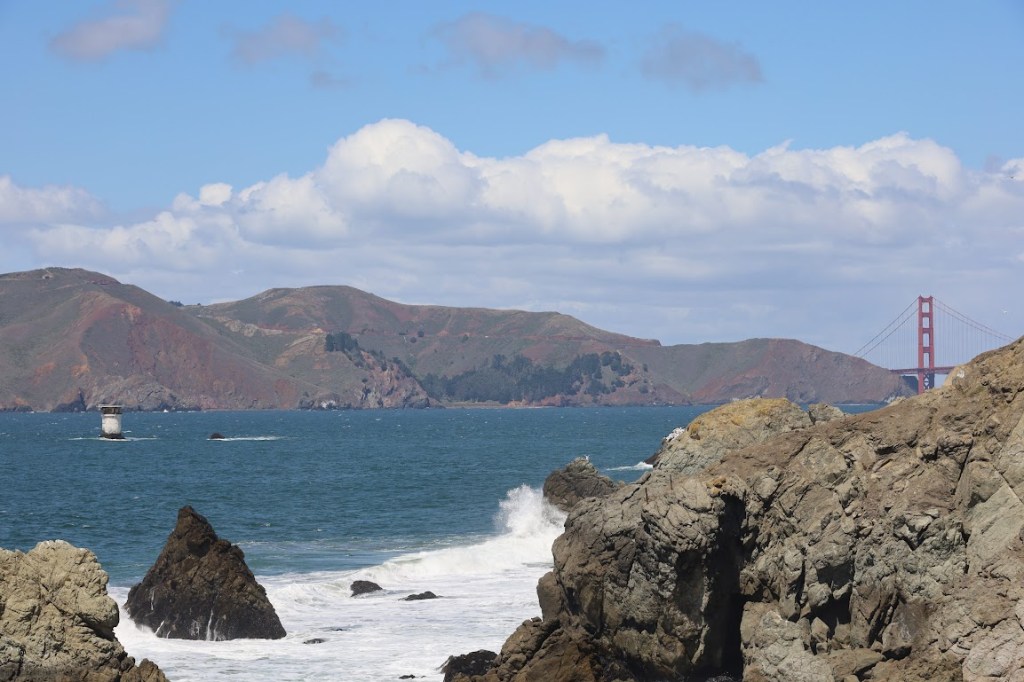 A photo taken from the top of a rocky cliff. More of the cliff is visible on the right, with waves crashing up against the rocks. A very small white structure is visible on the left in the distance. Behind that, brown hills mark the opposite shore. One of the Golden Gate Bridge's red towers can be seen in the distance on the right.