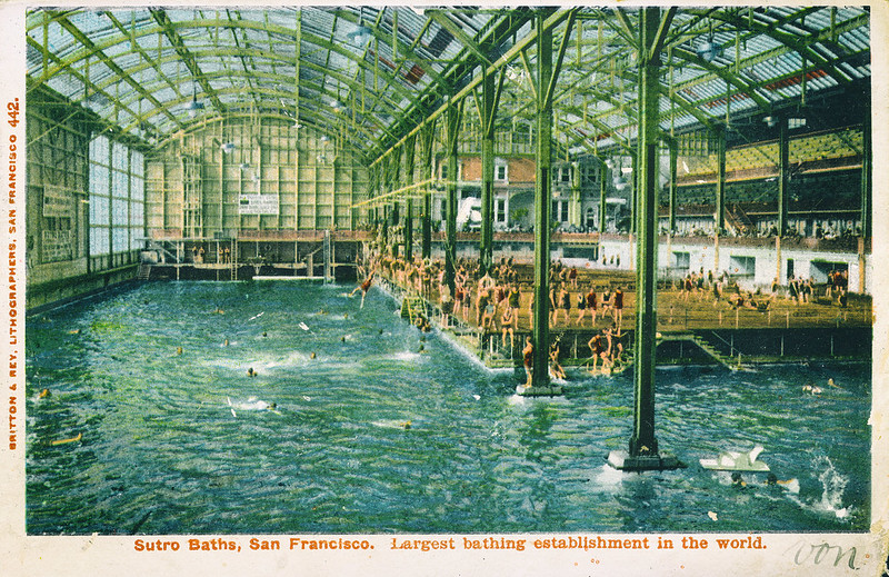 A vintage postcard reading, "Sutro Baths, San Francisco. Largest bathing establishment in the world." The image depicts the inside of a massive glass building, whose glass ceilings are held up by three-story metal posts. People in bathing suits mill around on a platform in the rear right quarter of the pool area. A few people swim in the pool. Their heads above the water appear as simple dots.