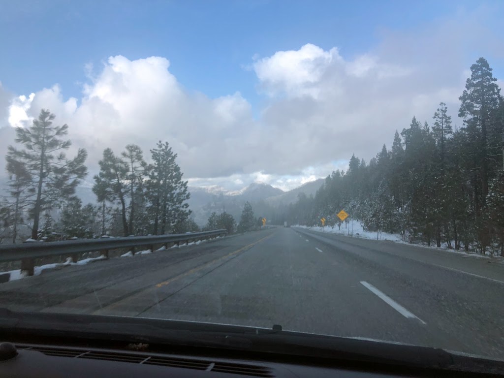A photo taken through the front windshield of the car as it drives uphill on a two-lane highway. In the distance, mountains are barely visible through what appears to be fog but is actually blowing snow. Dark clouds hover above the mountains, but blue sky is visible in the nearer distance.