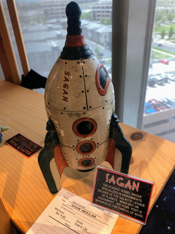 A cartoon-style sculpture of a rocket ship, resembling the sketch from the page above, except labeled "SAGAN." An auction label below the sculpture names the artist, Kyle Crutcher, and notes the minimum bid as $80 and the Quick Sale price as $135.