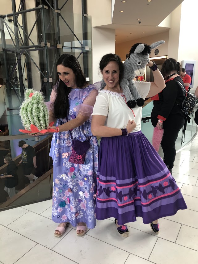Two women dressed in the costumes of Isabella and Luisa from the Disney movie Encanto. Isabella looks with delight at a large, crocheted cactus she holds in front of her, and Luisa flexes the muscles of one arm while carrying a large, crocheted donkey over her other shoulder. They look GREAT.