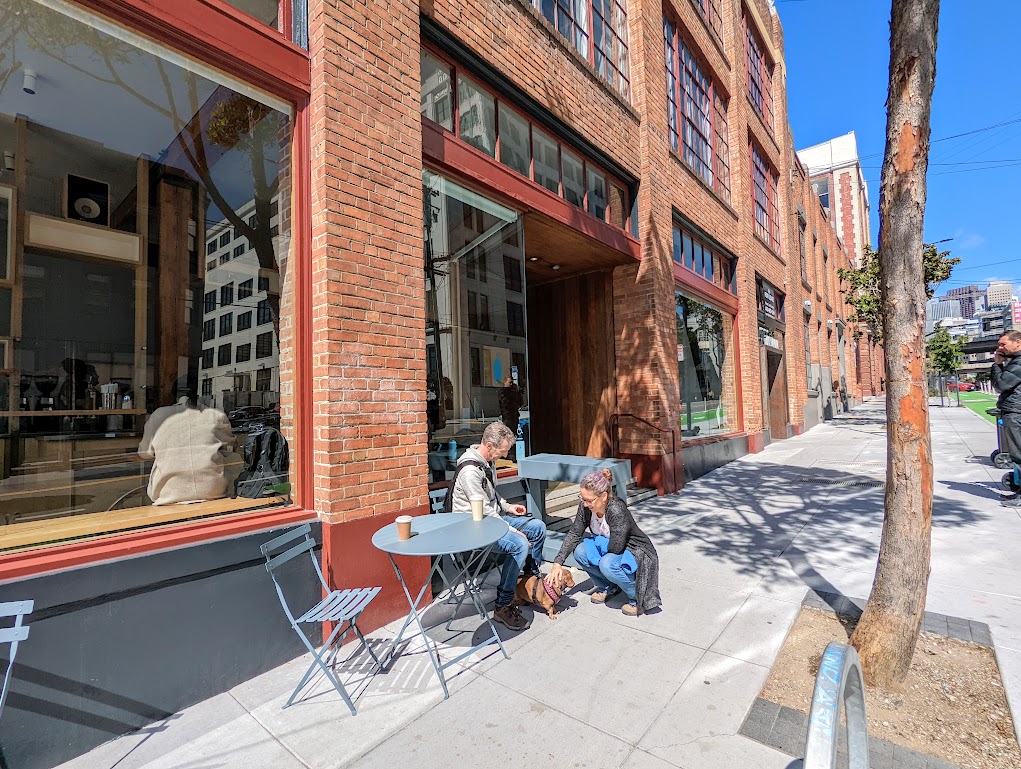 Matt sits at a small table on a sidewalk in front of a row of red brick shops. Gracie the dachshund is between his feet. Laura squats down and pets Gracie. There are two cups of coffee on the table. 