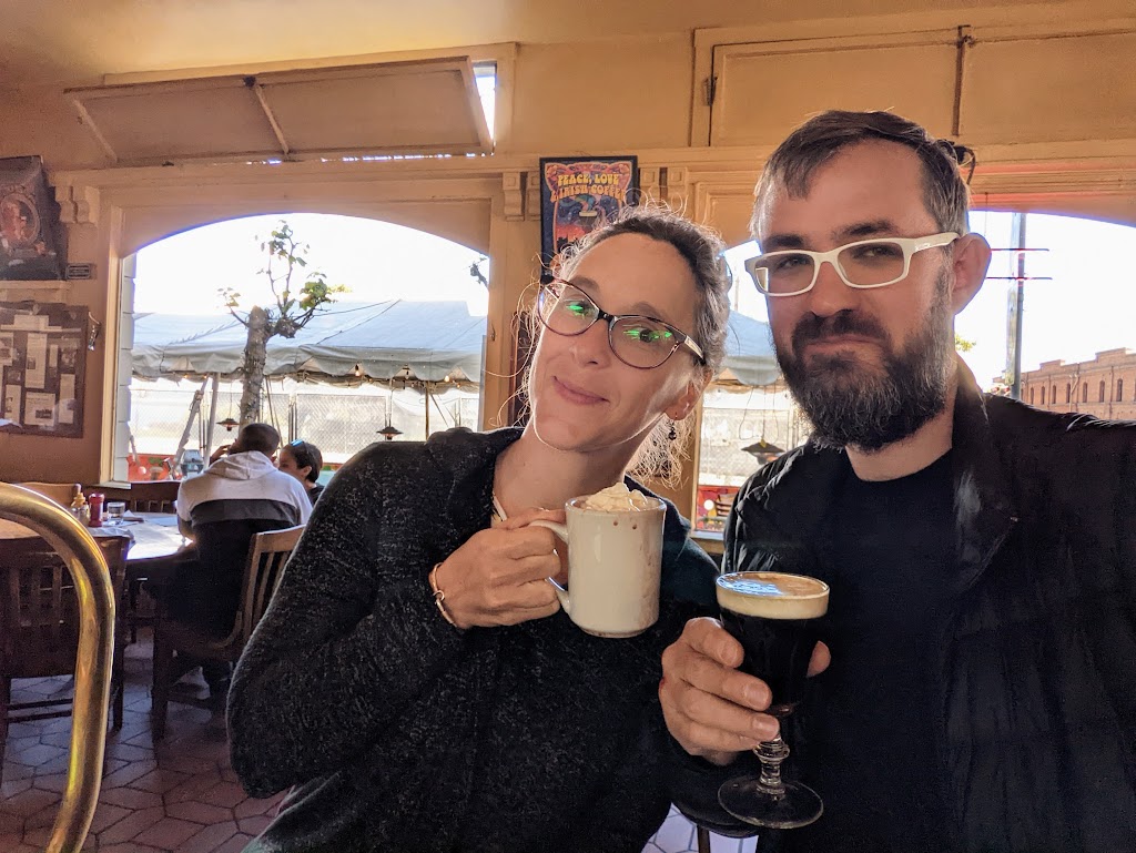 Laura and Dustin sit together inside a room with tables in the background. They lean together, each holding up a drink. Dustin's is in a glass goblet, black coffee topped with half an inch of irish cream. Laura's is a mug with dribbles of chocolate running down the outside. Both appear pleased with their beverages.