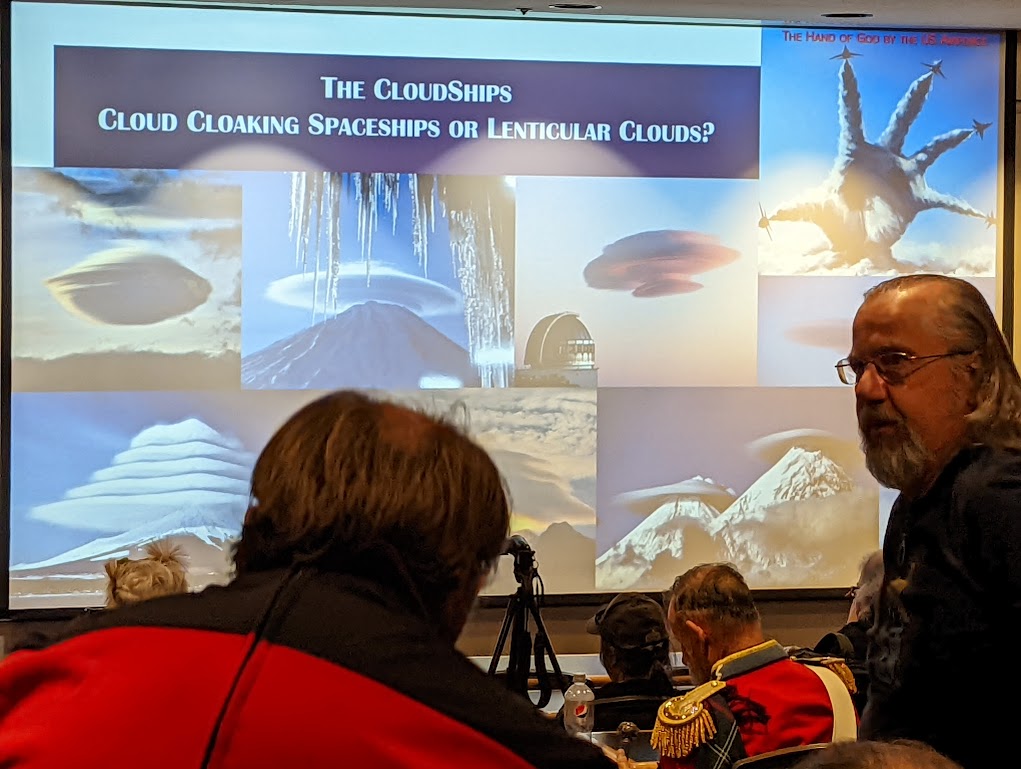 Photo taken through a crowd of people of a projection screen bearing the title "THE CLOUDSHIPS: CLOUD CLOAKING SPACESHIPS OR LENTICULAR CLOUDS?" Several photos of lenticular clouds that don't look much like spaceships at all fill the screen. 