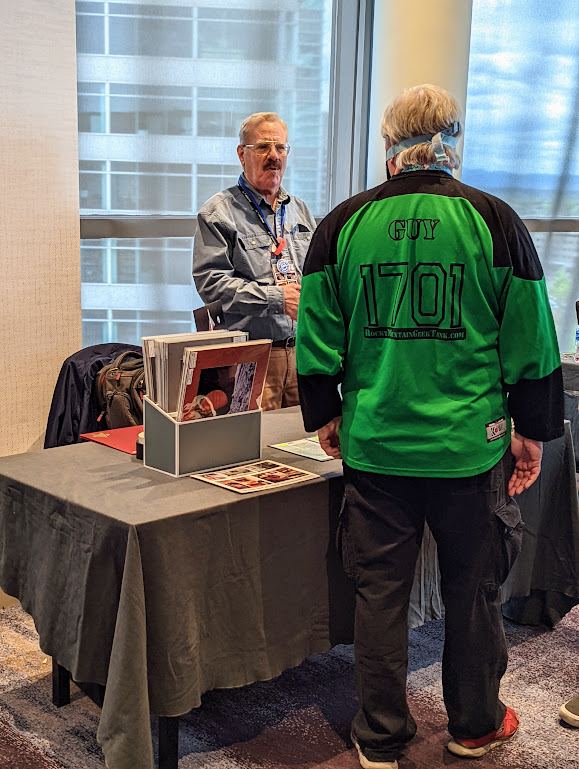 A man (a stranger) with his back to the camera converses with Rick Sternbach across a vendor's table. Several art prints are laid out on the table. 