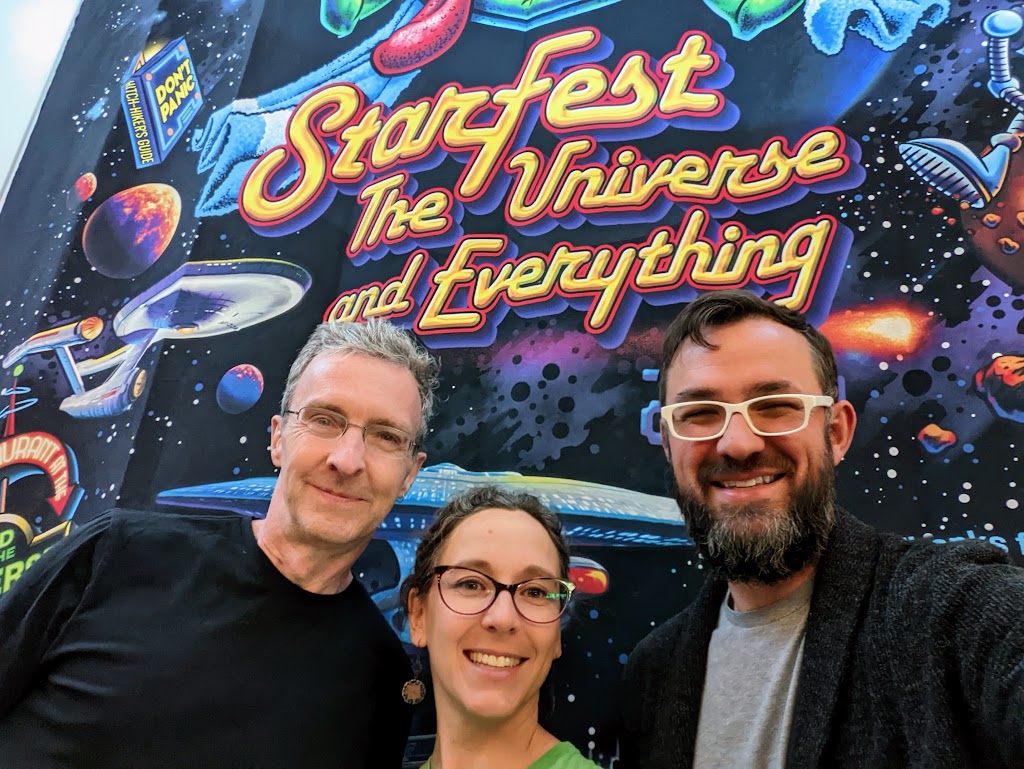 Matt, Laura, and Dustin pose in front of a large banner labeled "StarFest, the Universe, and Everything." The images on the banner are from Douglas Adam's Hitchhiker's Guide trilogy, including space ships, robots, planets, and neon signs.