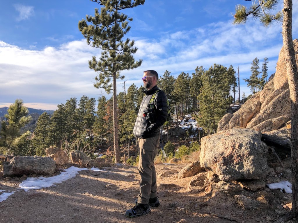 Dustin stands center, looking off to the left. He is on a dry, sandy patch with large granite boulders behind him and evergreen trees in the background. The sky is very blue with a few clouds on the horizon.