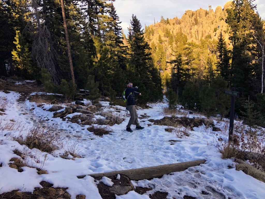 Dustin stands in the middle distance at a Y in the trail. He is pointing toward the right fork of the Y, looking back over his shoulder at the camera. The trails are covered with snow and surrounded by dark trees.