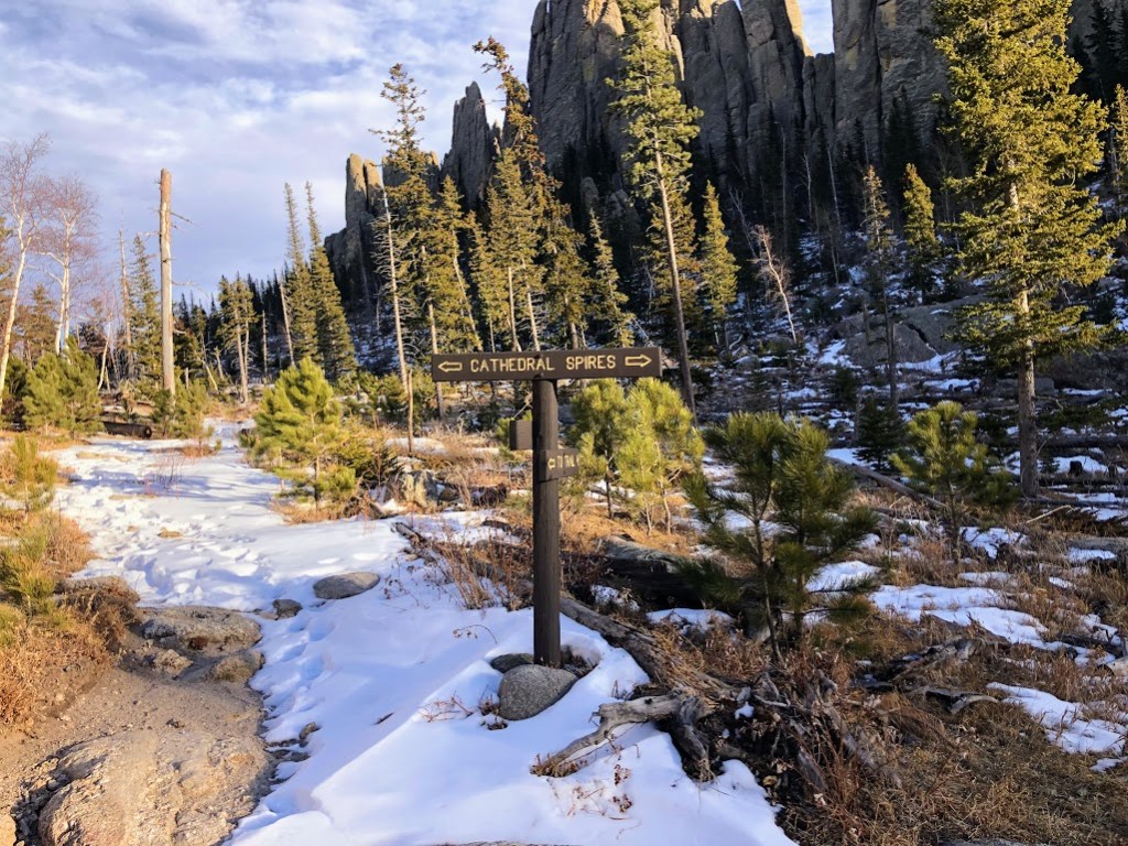 Looking along a snow-covered trail toward evergreens lit by the sun. A wood sign at center reads "CATHEDRAL SPIRES" and features arrows pointing to both sides. 