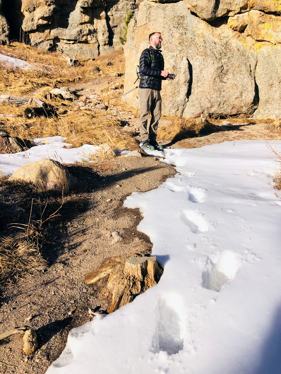 Dustin stands center, in front of a large granite wall, looking toward the right side of the photo. He has left a row of footprints in the snow along the trail which show deep shadows because the snow is very deep. Gravel beside the snow suggests the snow should not have been so deep.