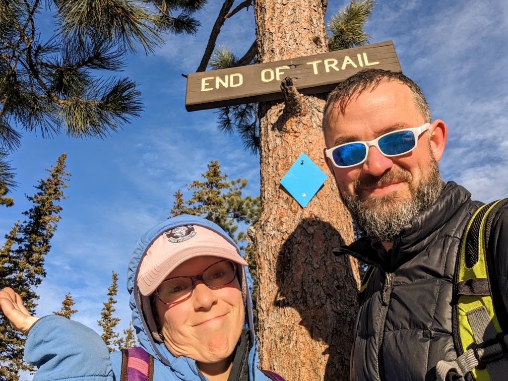 A selfie of Laura and Dustin standing in front of a tree bearing a sign that reads "END OF TRAIL". Laura holds up a hand in a shrug to say "oh well," Dustin smiles like a normal person. 