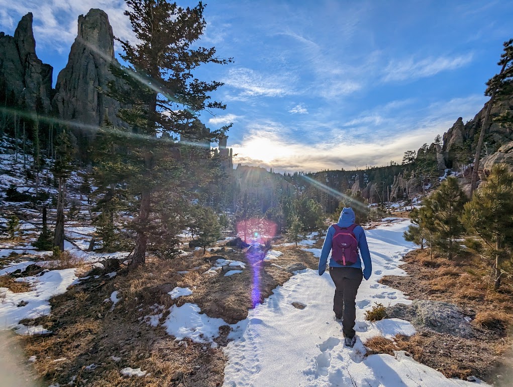 Laura walks away from the camera along a snow-covered trail toward a sun that sits low on the horizon. To both sides the trail is lined with pine trees of many sizes. A very tall granite spire rises in the distance on the left.