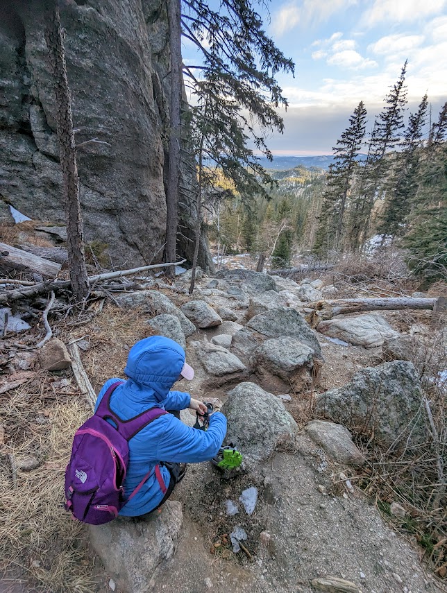 Laura sits at the top of a steep, boulder-covered slope, removing her microspikes. There is no snow anywhere to be seen.