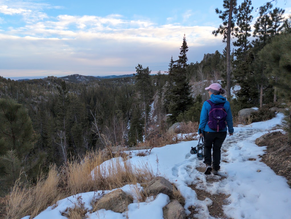 Laura walks along a snowy trail with many footprints tracked into it, carrying her microspikes. She looks over her left shoulder out at a view of dark granite and trees.