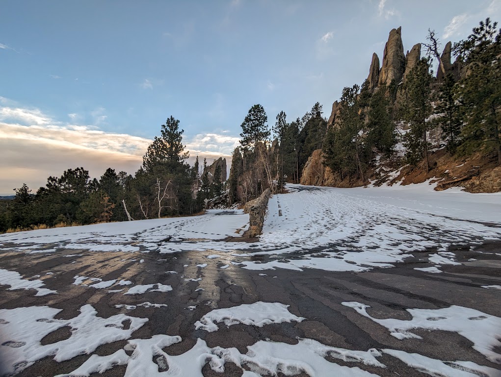 A photo of a hairpin turn in a road, taken from the bend of the pin. On the left, the road leads away from the camera going downhill. On the right, the road leads away from the camera going uphill. The road is covered with snow and footprints.