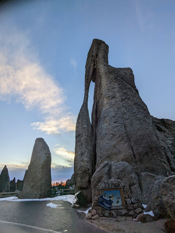 A granite spire that looks a whole heck of a lot more like a needle's eye than that tunnel did. The spire appears to be 50 or 60 feet tall. An interpretive sign can be seen at its base but is too small to read. In the background, a stripe of orange sunset shows on the horizon.