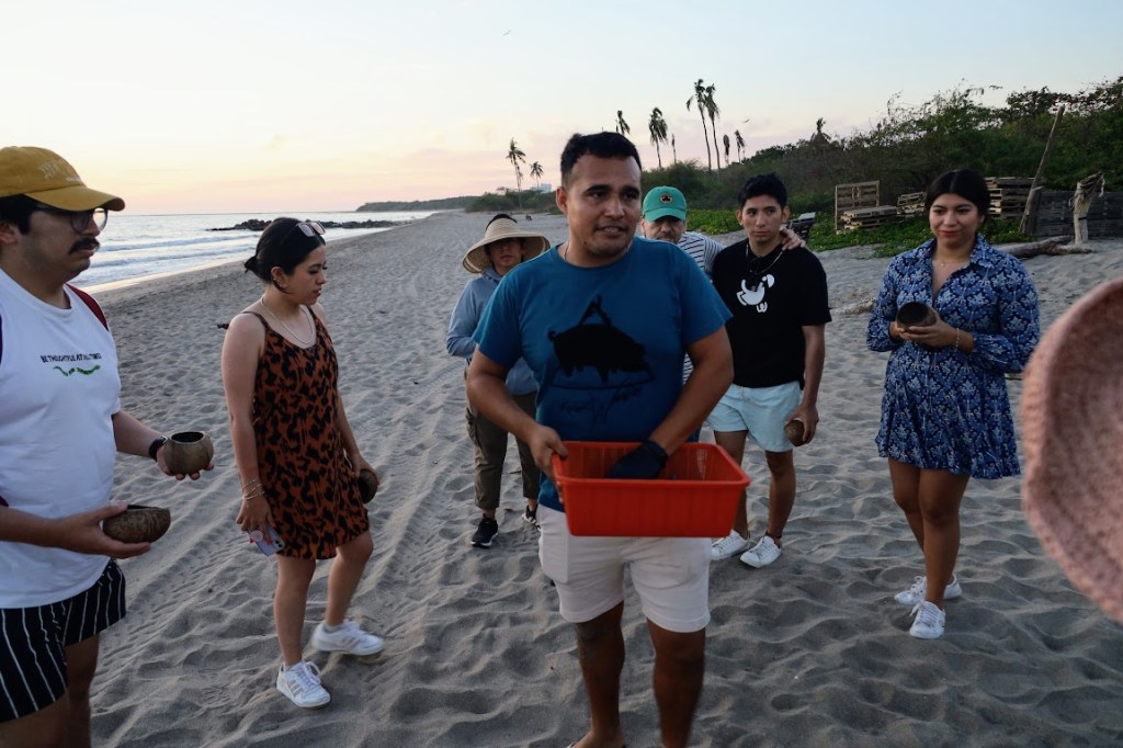 A man in a blue shirt stands at center, with a ring of people behind him and presumably stretching to include the photographer. The man holes an orange, mesh, plastic basket in front of him. The people standing around him are holding coconut shells. The ocean can be seen in the background to the left. 