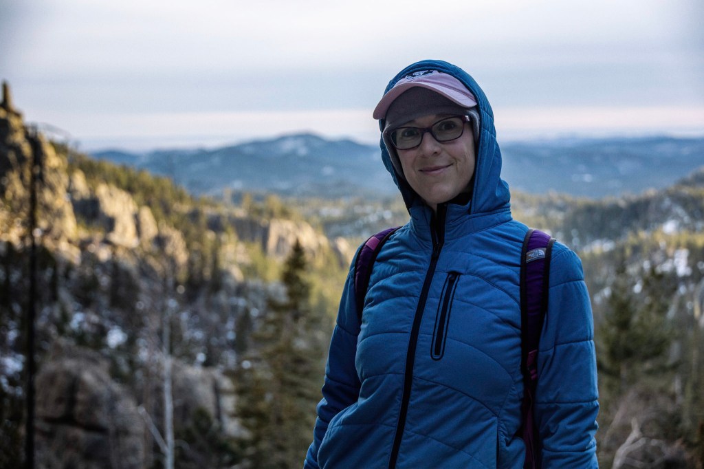A portrait of Laura wearing a head band covered by a baseball cap covered by the hood of her jacket. She looks warm. The background of Black Hills granite and pine trees is out of focus.