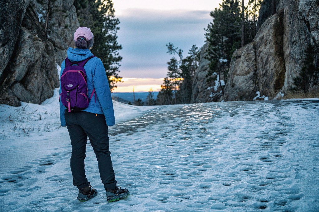 Laura stands with her back to the camera on a paved road that has been covered in snow that is now well trampled by many feet. Granite walls line both sides of the road, but in the distance beyond the road, orange bands of sunset can be seen. The light glints off ice under patches of snow.