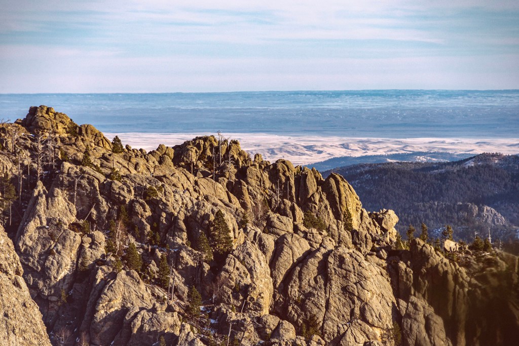 An entire hillside composed of weathered granite lit by the sun. In the background a dark tree-covered slope, and beyond that the flatter land of the prairies stretches into the distance.