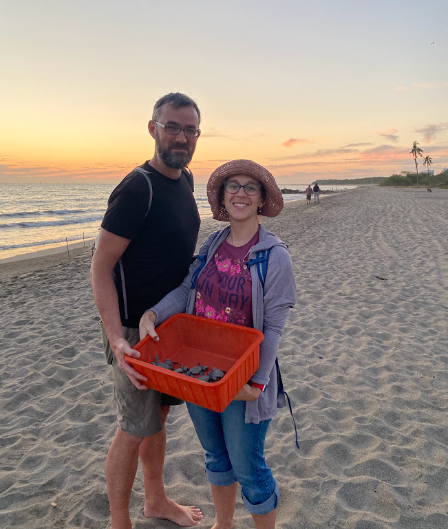 Dustin and Laura stand on a sandy beach with the ocean in the background and the sky showing sunset colors. They are holding the plastic orange basket from the previous photo, which you can now see contains baby sea turtles. 