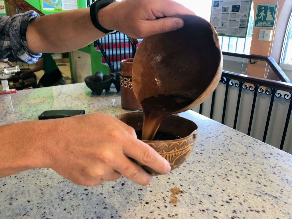 A closeup of Dustin's hands, each holding half a jicara shell, pouring a dark brown liquid from one to the other.