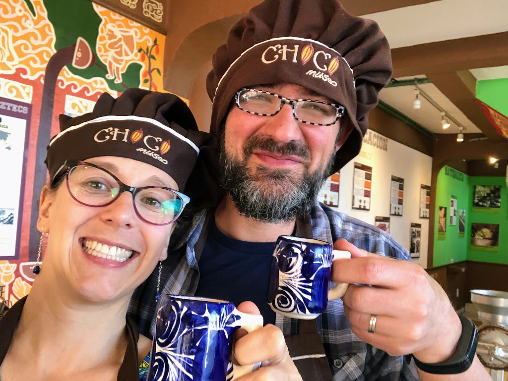 A selfie of Laura and Dustin, grinning at the camera, wearing brown "chef" hats that say "ChocoMuseo," holding up blue and white painted mugs.