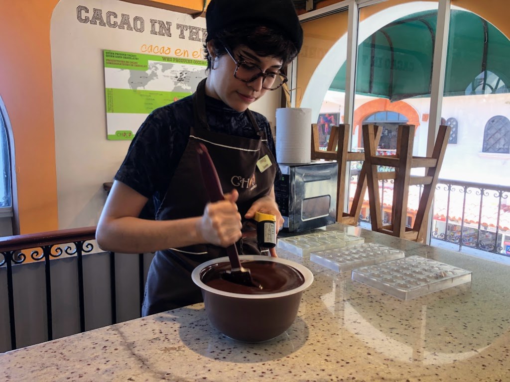 Lizzette stirs a large bowl full to the brim with dark chocolate. She holds the spatula in one hand and a remote thermometer pointed at the chocolate in the other.