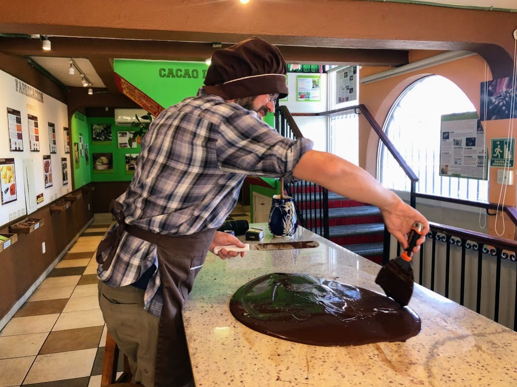 Dustin leans over the counter, holding a putty knife in one hand and a flat metal spatula in the other. He is using the putty knife to scrape one edge of a puddle of dark chocolate toward himself. The chocolate looks like it's trying to escape, because it is.