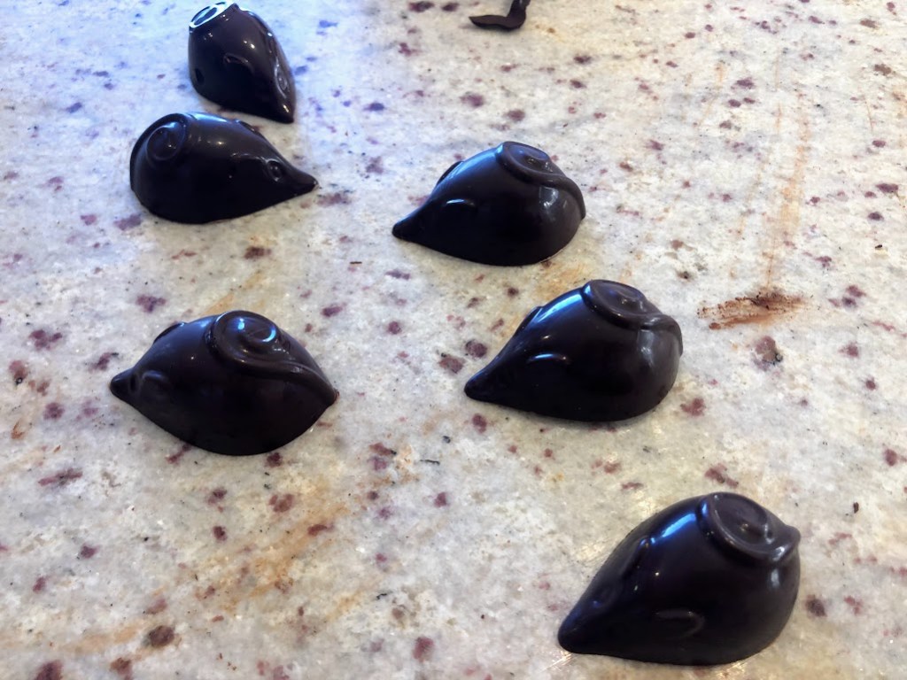 A closeup of teardrop-shaped chocolate mice with very pointy noses and little tails curled into a spiral on their backs. They are very dark chocolate, and very shiny.
