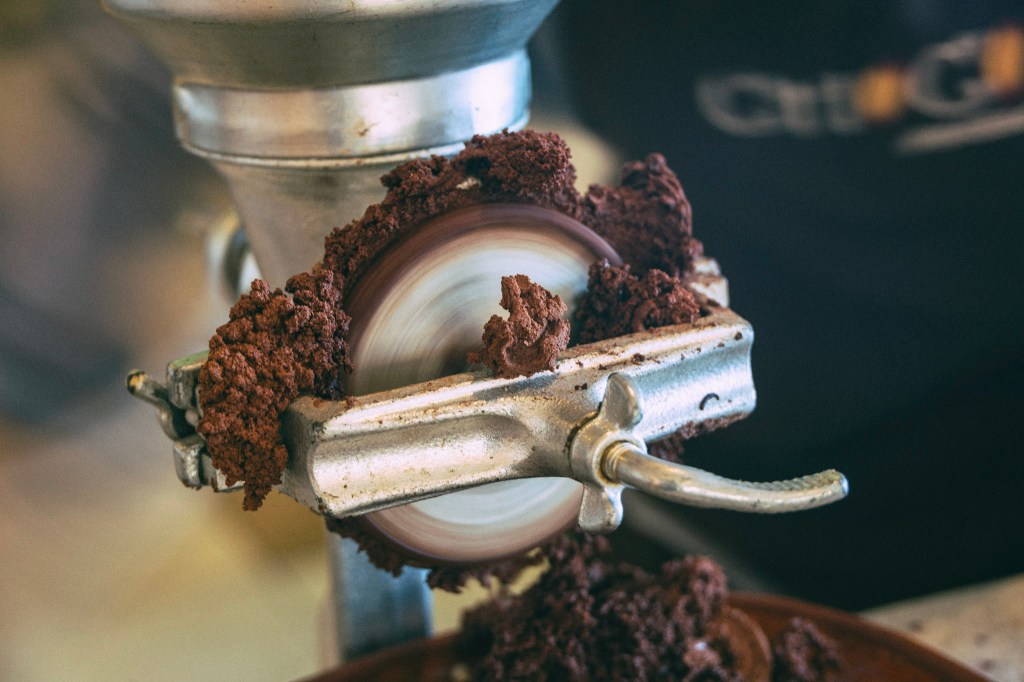 Closeup of a grinding wheel with coarse, dark brown cacao paste extruding from the edges. The wheel is blurred with the fast motion of its spin.