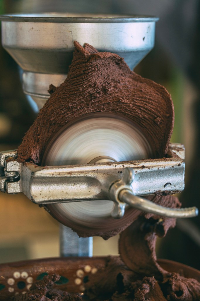 Closeup of a grinding wheel velvety-looking cacao paste and sugar, now slightly lighter brown in color, extruding from the edges. The wheel is blurred with the fast motion of its spin.