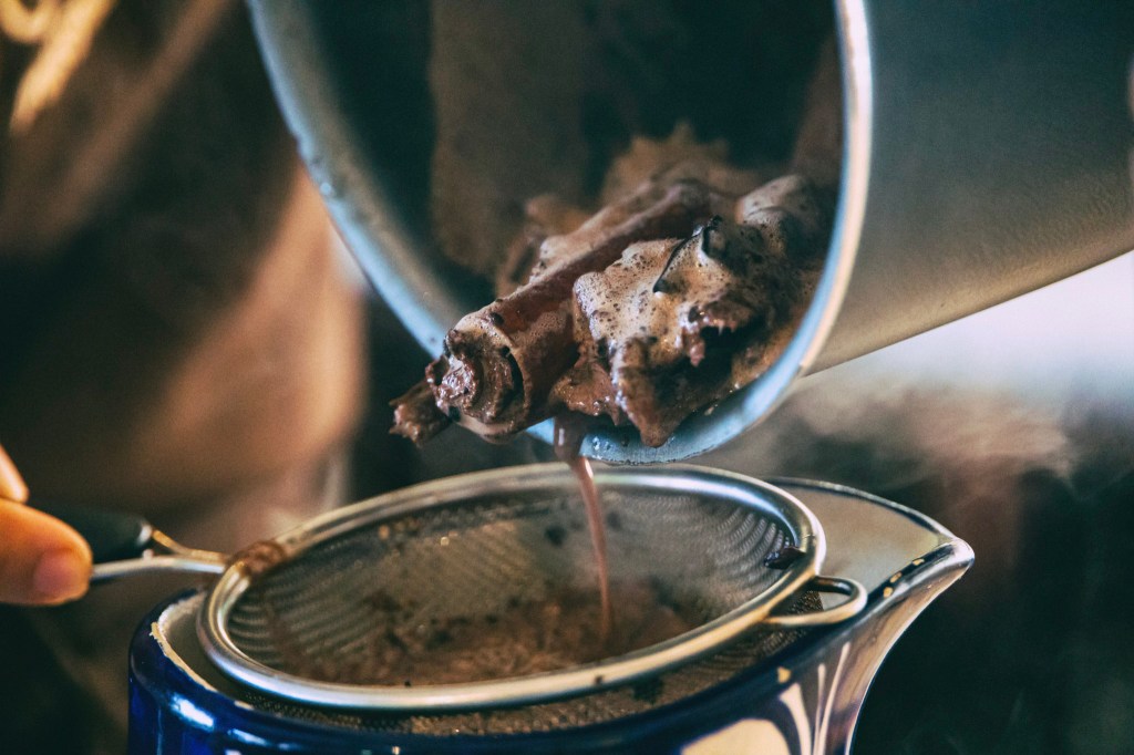 A closeup of liquid being poured from a pot through a strainer into a blue-and-white painted pitcher. Cinnamon sticks, cloves, and other debris hang at the edge of the pot lip. The liquid coming through looks like - and is! - hot chocolate.