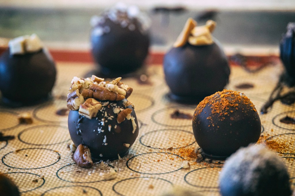 A closeup of a tray of dipped dark chocolate truffles with various toppings - pecans, chili powder, peanuts, coconut, tejate, ginger. They look impressively like real truffles.