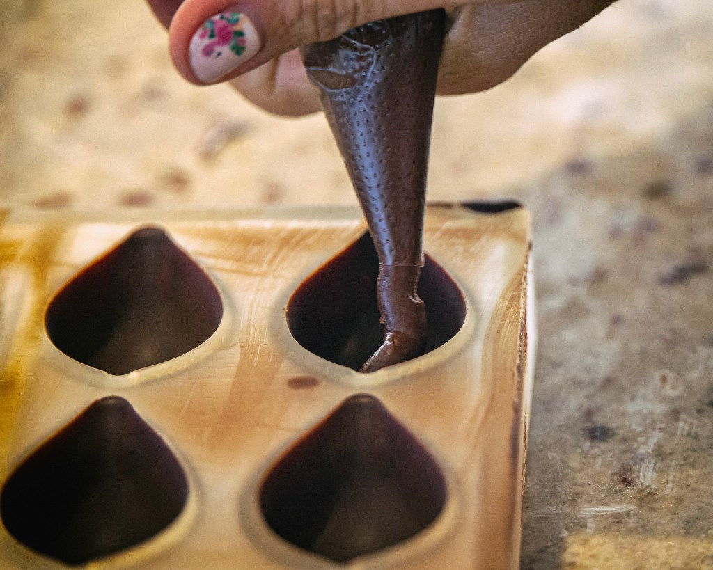 Closeup of a corner of a polycarbonate mold whose four teardrop shapes have been coated with chocolate. Laura's hand holds a plastic piping bag full of ganache, which she is squeezing into the top-right shape.