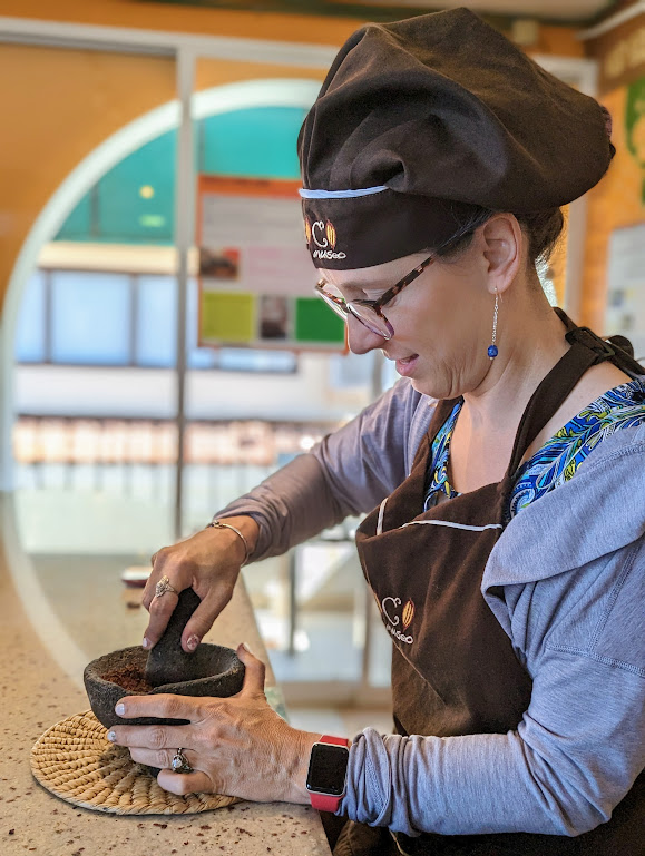 Laura, in profile, uses a black mortar and pestle to grind a dark brown paste. She appears amused and a little skeptical.