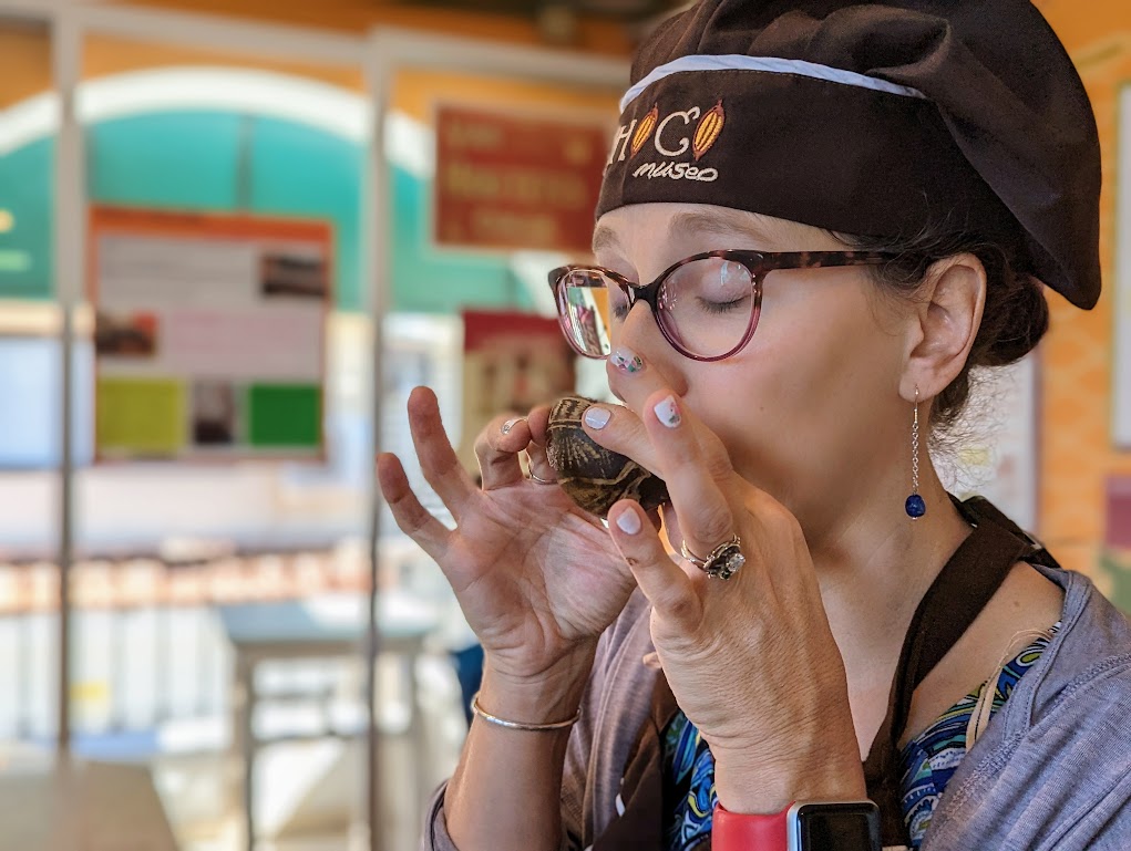 A portrait of Laura, wearing her ChocoMuseo hat, eyes closed, sipping out of a tiny jicara shell with carvings on the outside. Her pinkies are up like a fancy person.