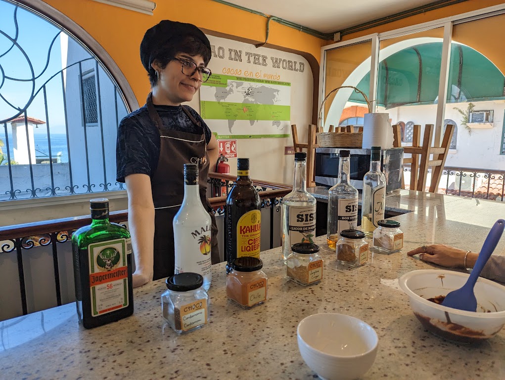 Lizzette stands behind the counter, one hand on her hip, looking proud. Lined up on the counter in front of her are bottles of Jagermeister, Malibu, Kahlua, orange liqueur, spiced rum, and tequila. Jars containing various spices are lined up in front of the booze bottles. 